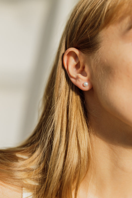 Natural 6mm pearl stud earrings on a woman’s earlobes, featuring lustrous white pearls and 925 sterling silver posts with a rhodium-plated finish